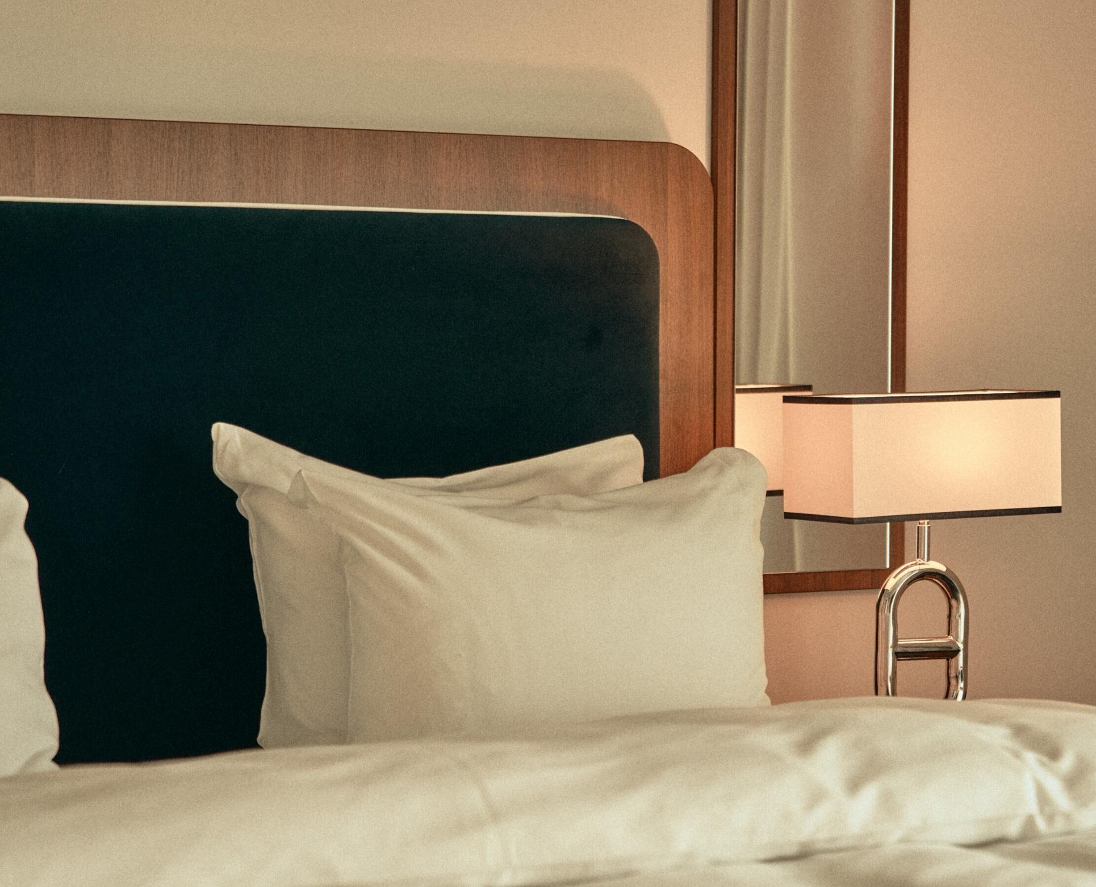 Una cama bien tendida con sábanas blancas, cabecera oscura y lámpara de noche con pantalla rectangular.