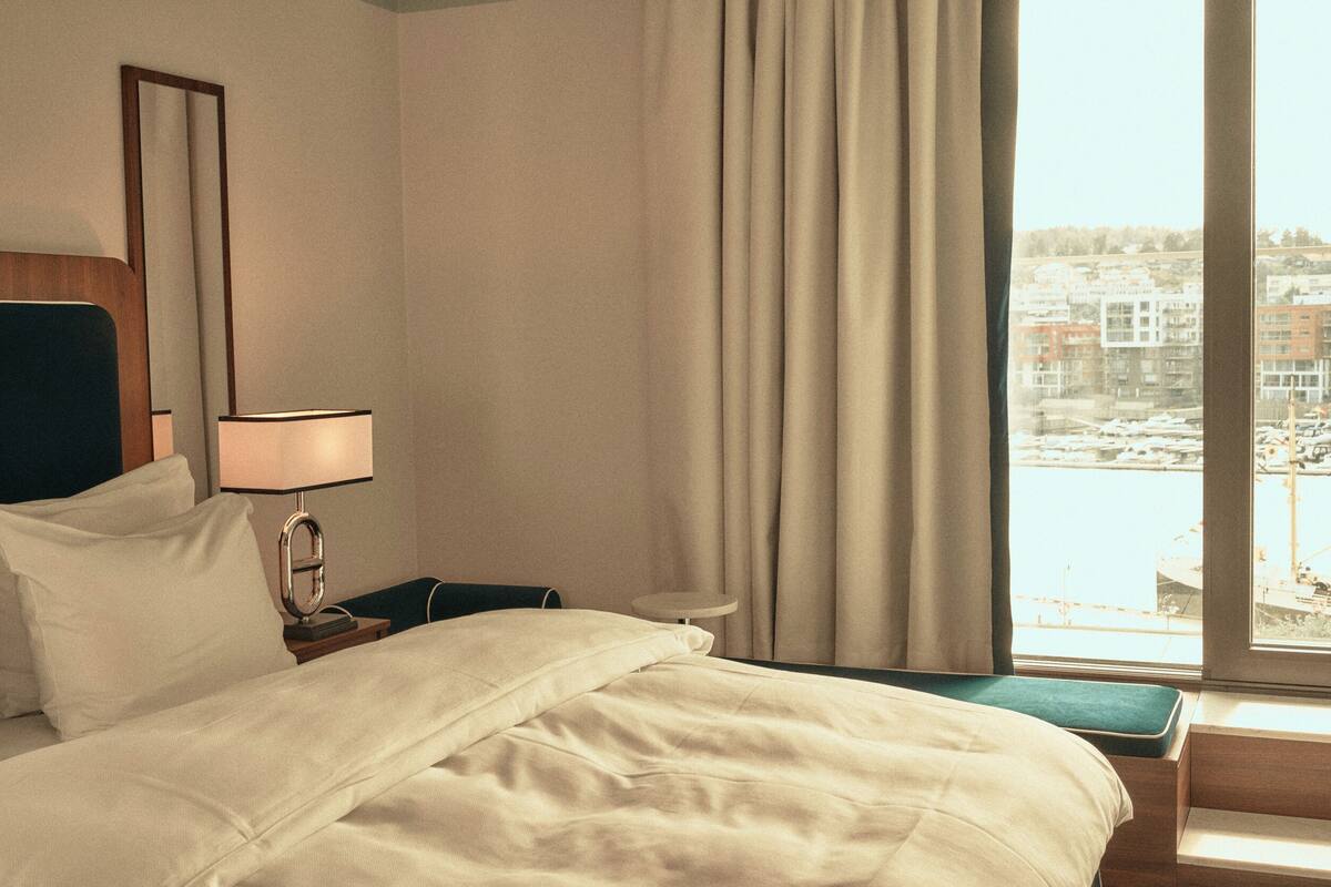 A hotel room with a bed, a nightstand with a lamp, a view of a cityscape through a window with curtains.
