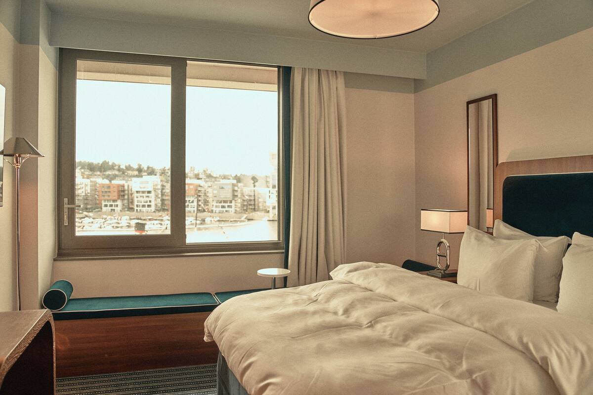 A hotel room with a large window, a bed, a bedside table, and a lamp.