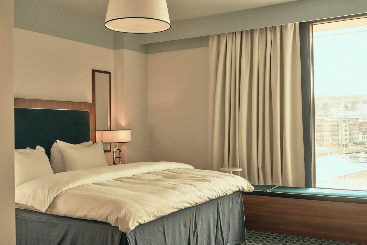 A hotel room with a bed, a bedside lamp, a window with curtains, and a view of buildings outside.
