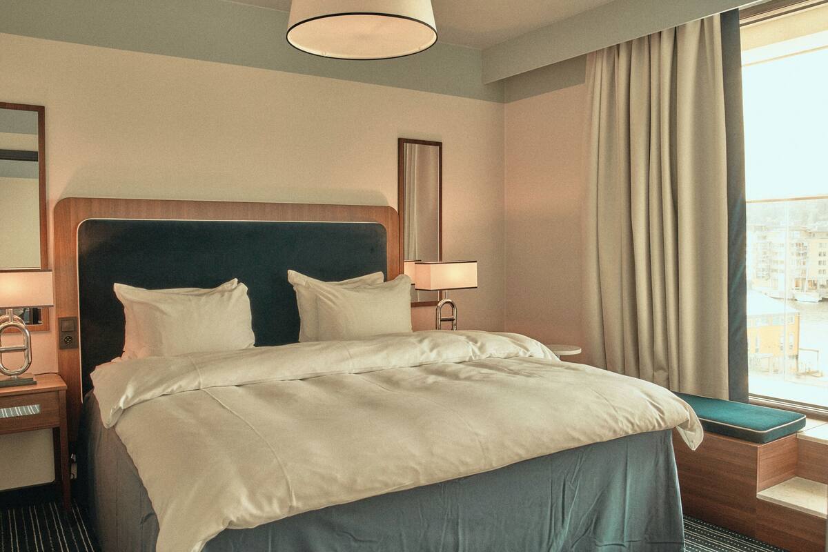 A hotel room with a large bed, a bedside table with a lamp, a window with curtains, and a view of buildings outside.