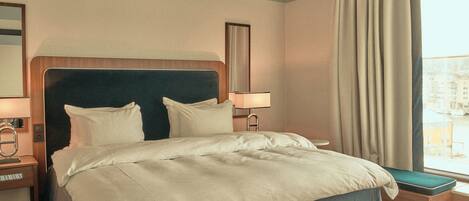 Habitación de hotel con una cama grande, una mesita de noche con lámpara, una ventana con cortinas y vistas a edificios.