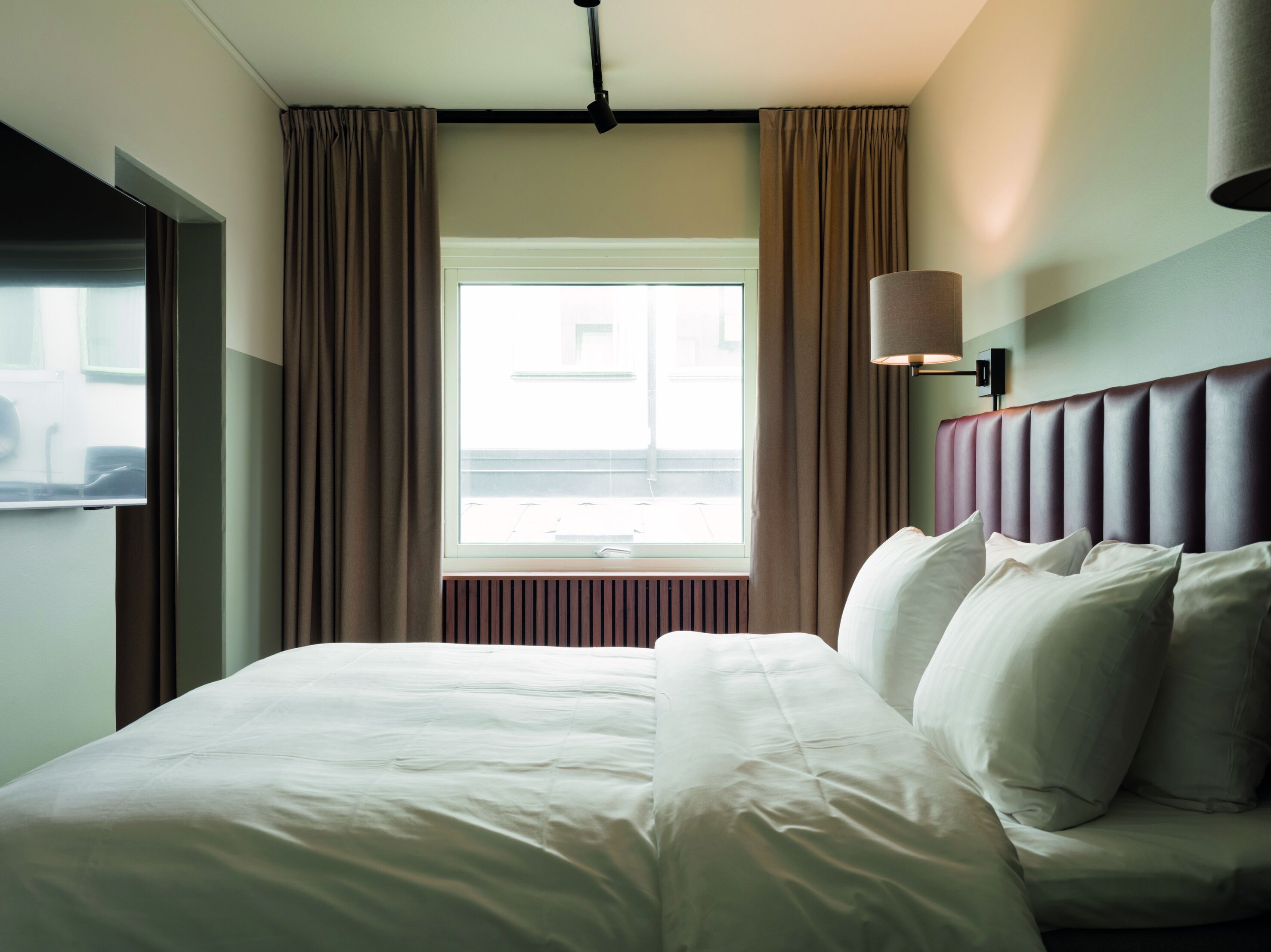 A hotel room with a large bed, a window with curtains, a wall-mounted lamp, and a radiator.