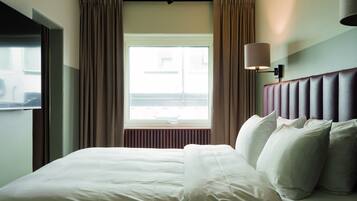 A hotel room with a large bed, a window with curtains, a wall-mounted lamp, and a radiator.