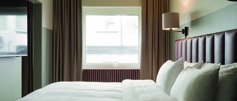 A hotel room with a large bed, a window with curtains, a wall-mounted lamp, and a radiator.