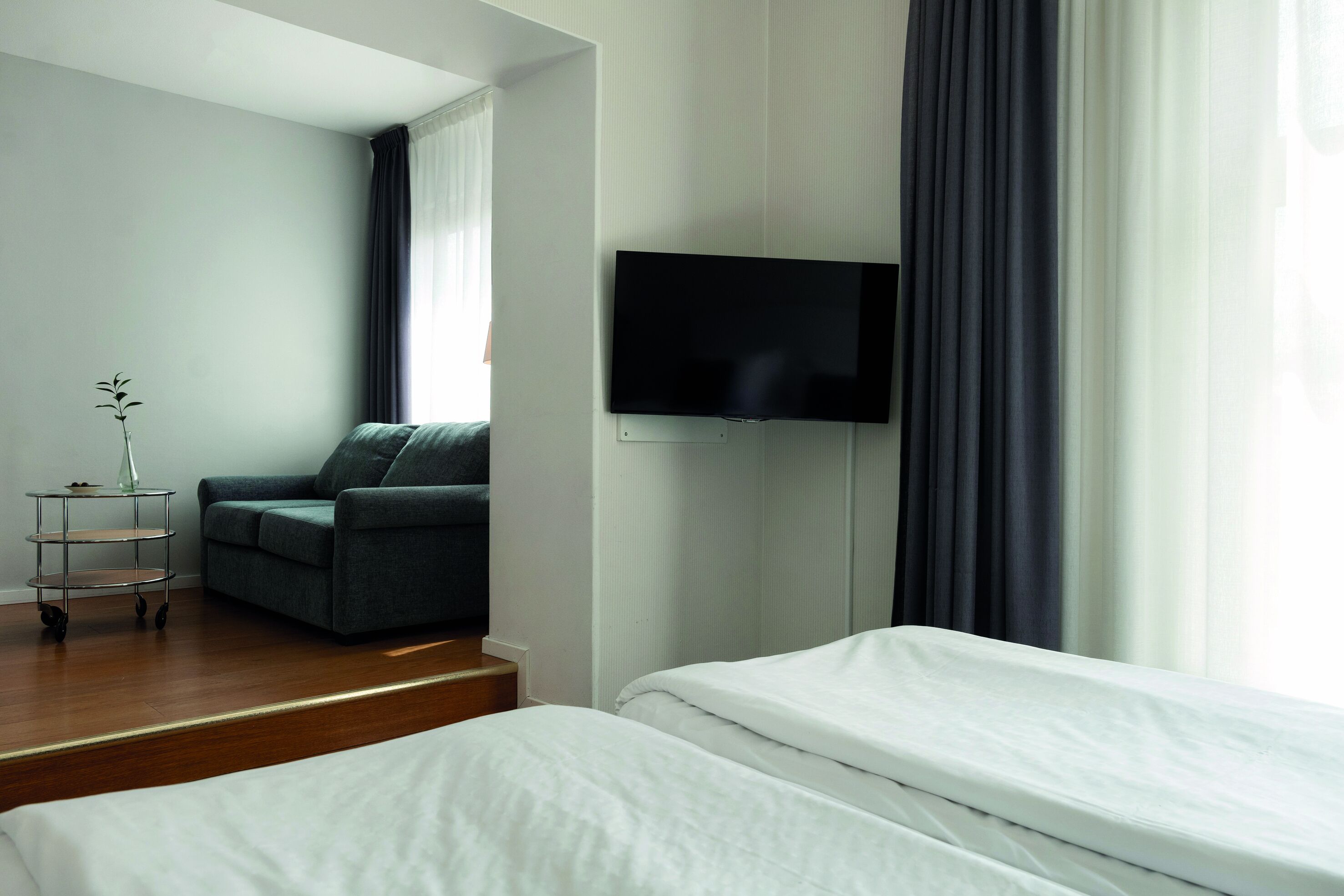 Quarto de hotel com cama, sofá, televisão fixada na parede e uma pequena mesa com um vaso.