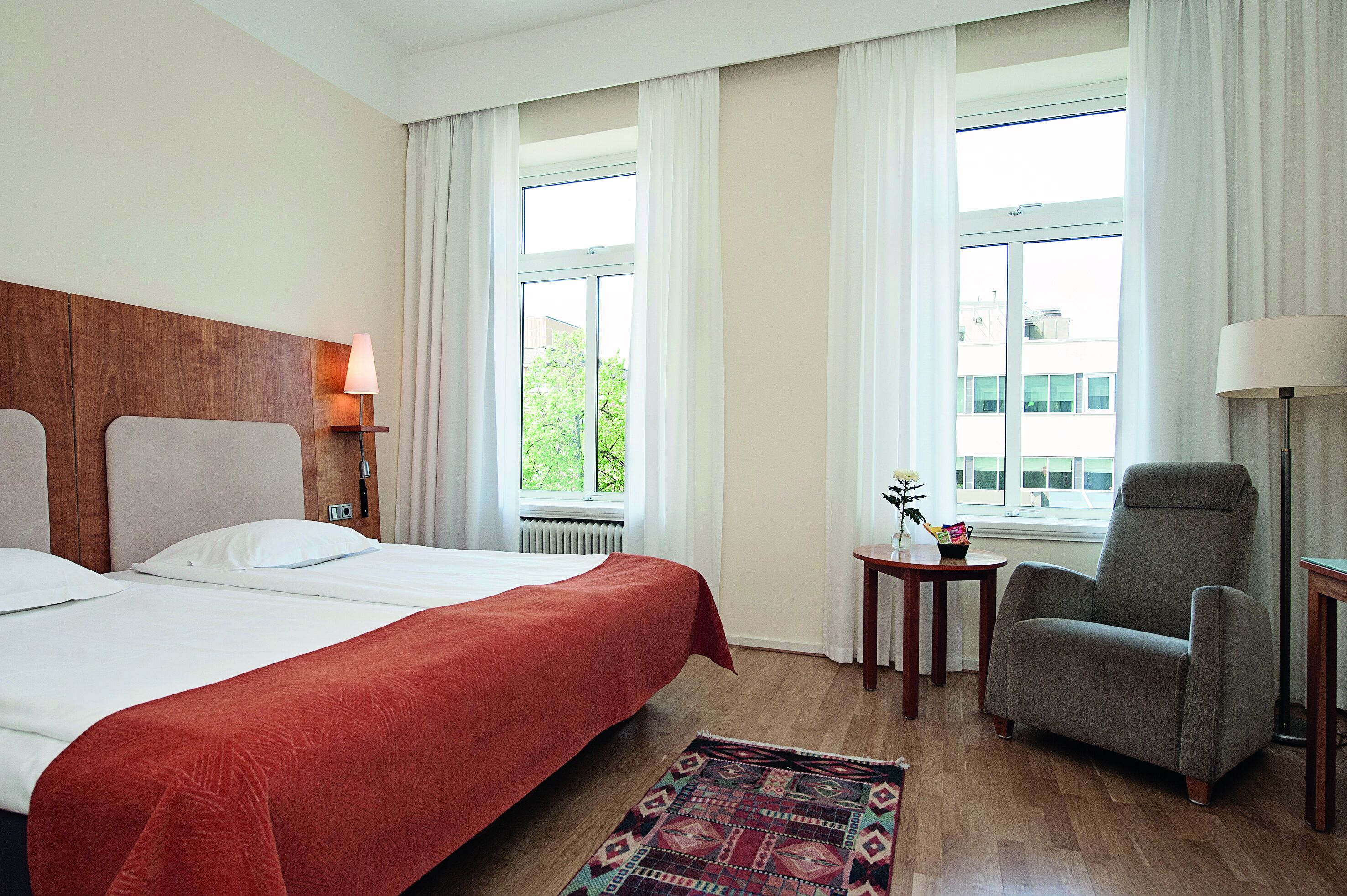 A hotel room with a large bed, a chair, a small table, and a window with curtains.