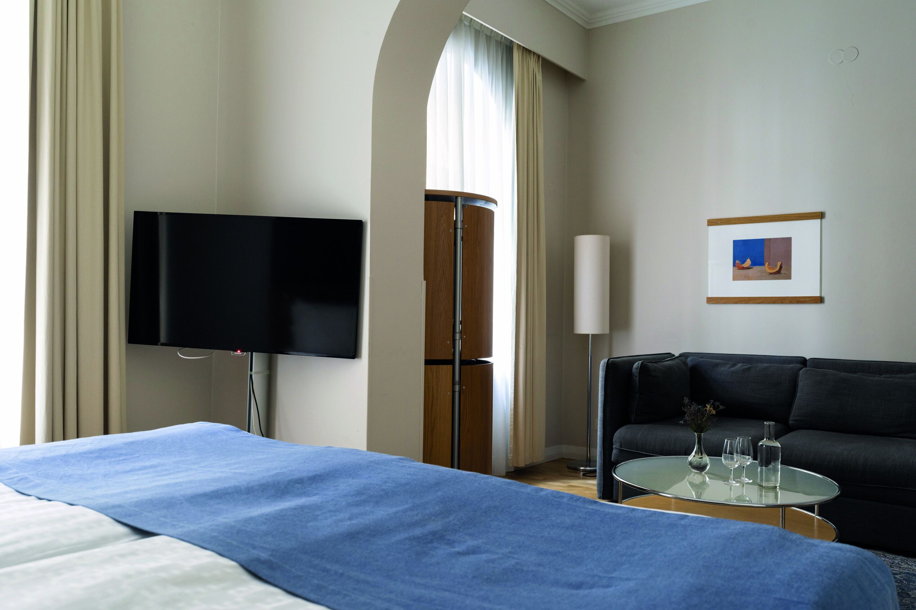 Una habitación de hotel con cama, televisor, sofá, una mesita con vasos y un cuadro enmarcado en la pared.