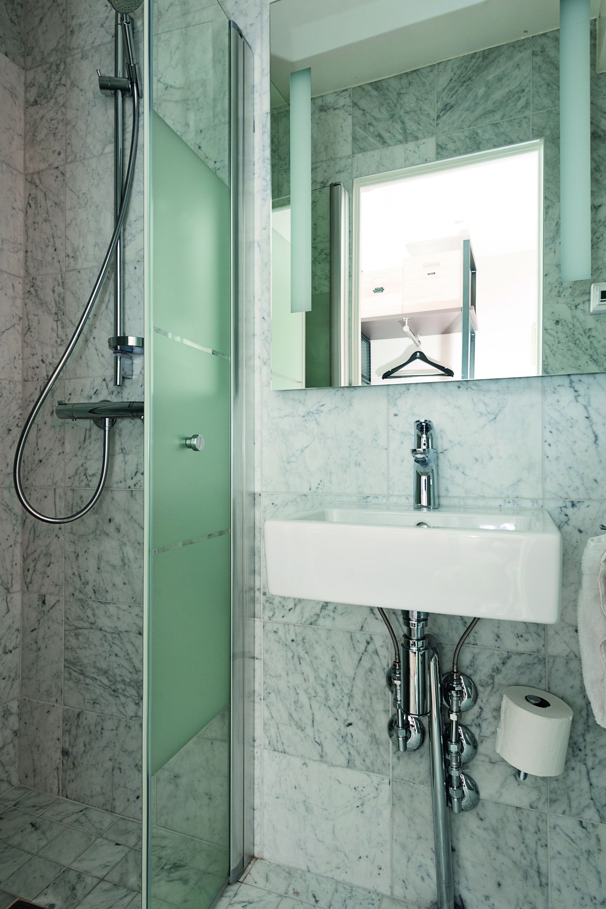 Un baño con ducha separada por vidrio, lavamanos blanco y pared con azulejos de mármol.