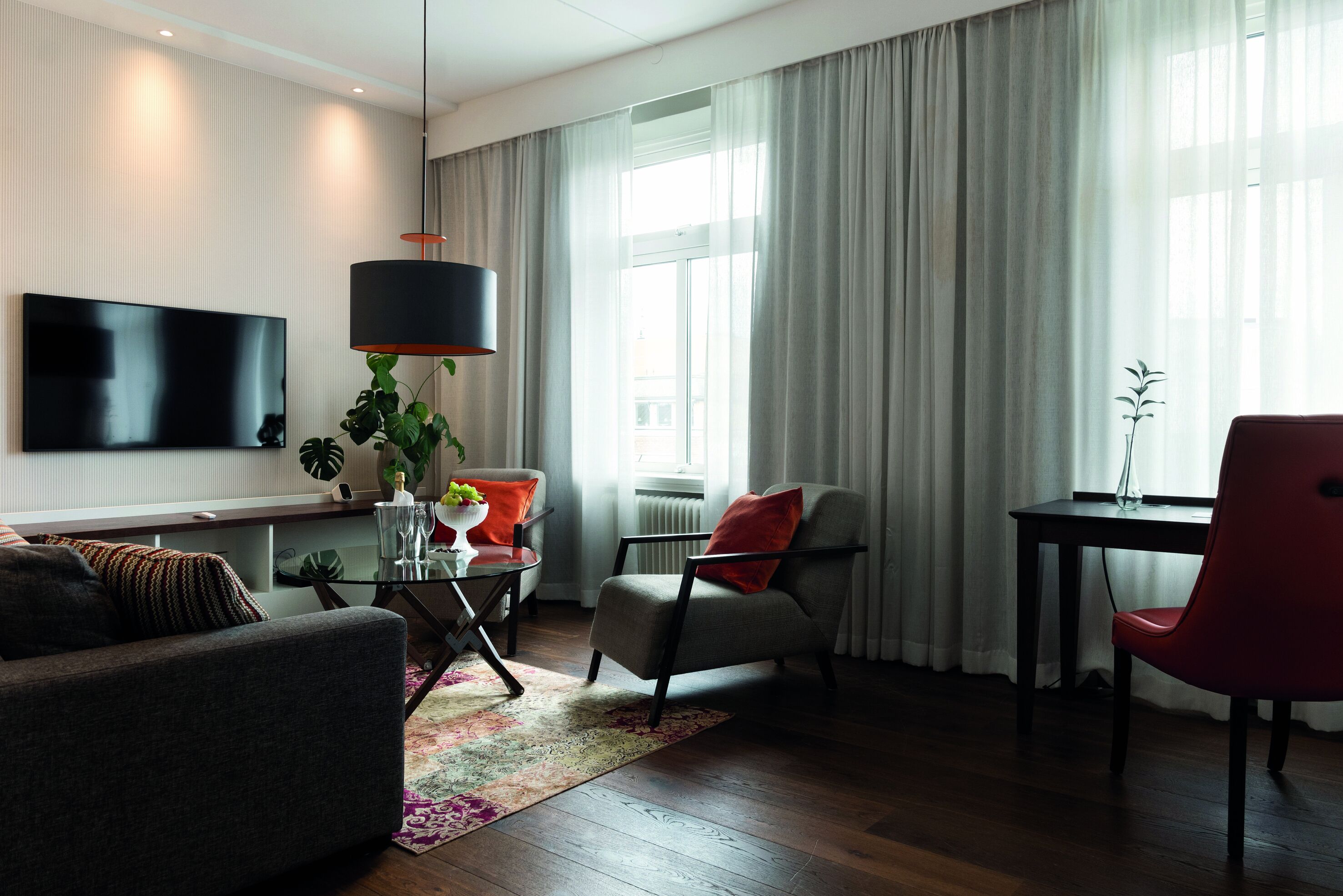 Una sala moderna con un televisor de pantalla plana, una mesa de centro de vidrio, un sofá, una silla roja y una mesa de comedor con un jarrón.
