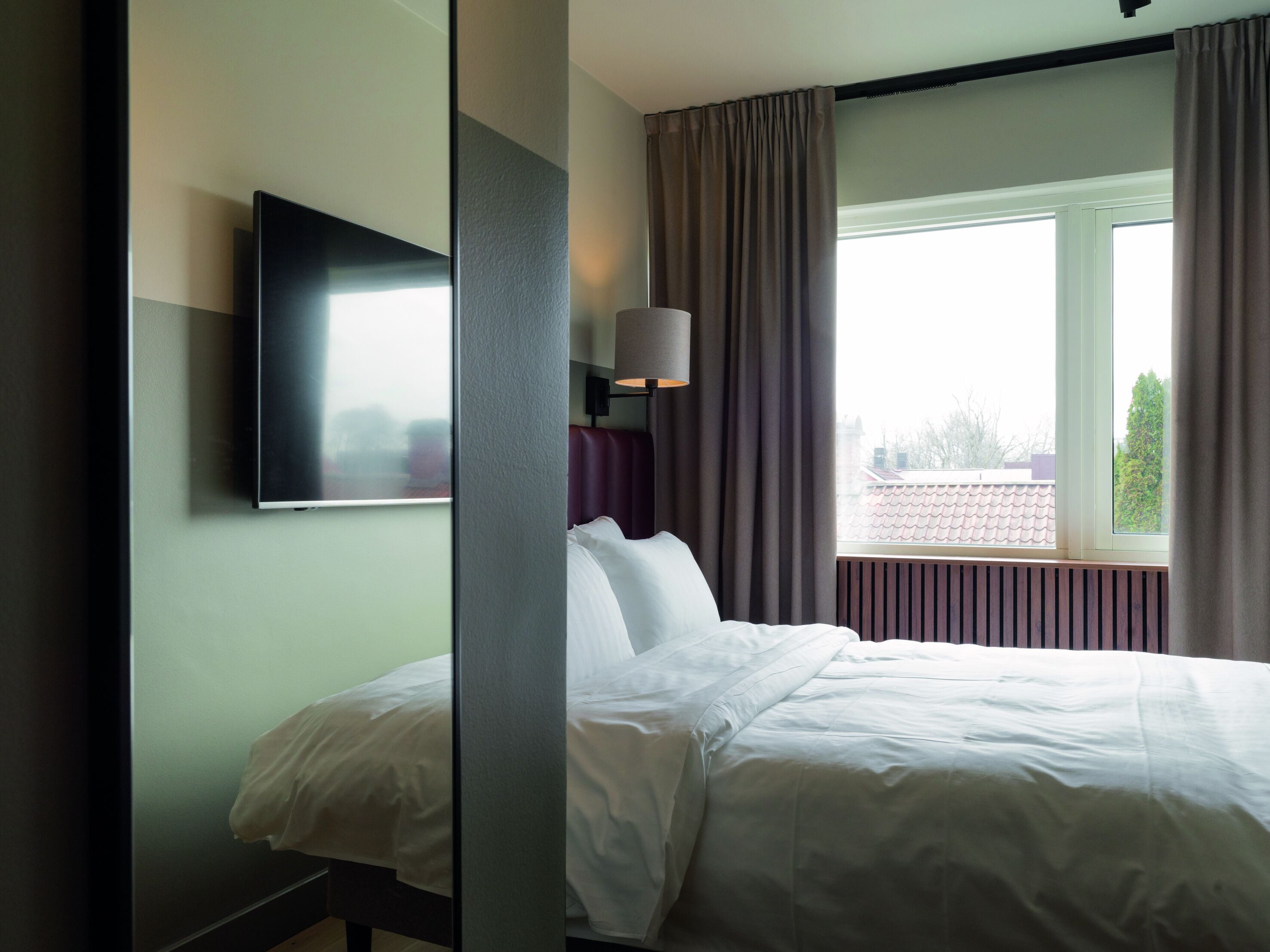 A hotel room with a bed, a flat-screen TV mounted on the wall, a window with curtains, and a lamp on a bedside table.
