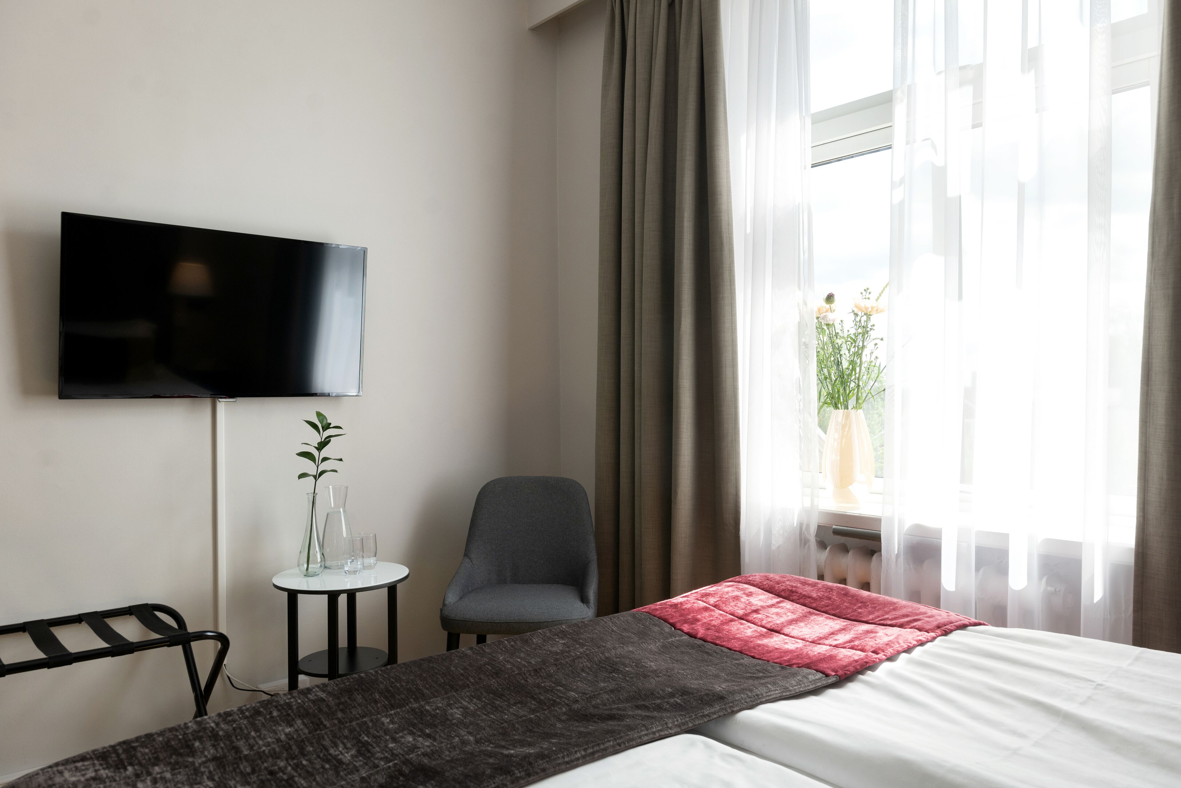 Habitación de hotel con cama, televisión montada en la pared, silla, mesita con un jarrón y vasos, y ventana con cortinas.