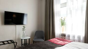 Habitación de hotel con cama, televisión montada en la pared, silla, mesita con un jarrón y vasos, y ventana con cortinas.