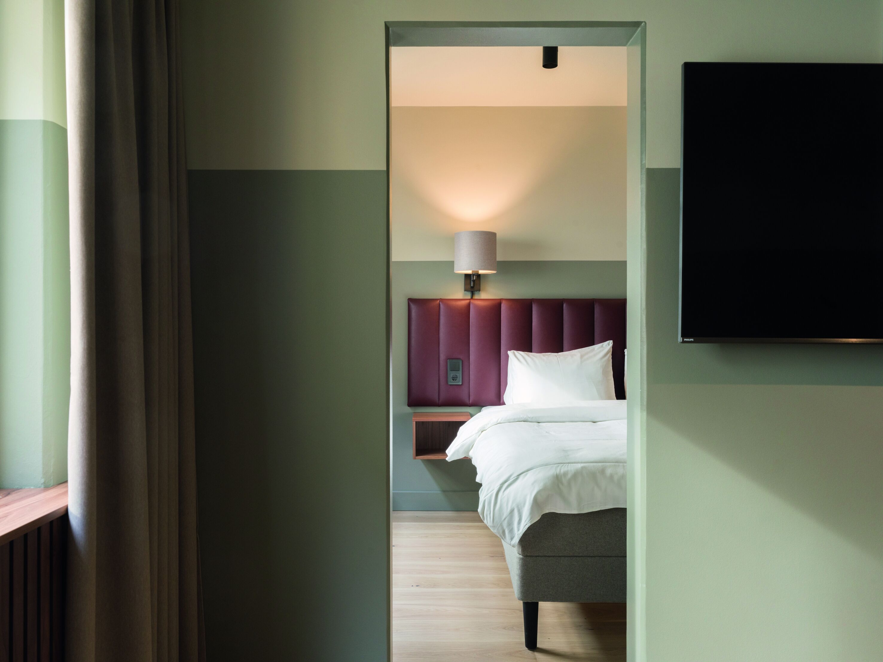 Habitación de hotel con cama, mesita de noche, lámpara y televisor montado en la pared.