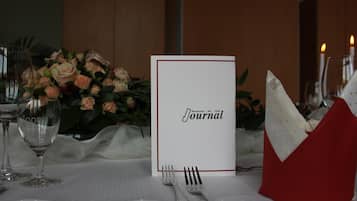 Ein Tisch ist mit einer weißen Tischdecke, einer Speisekarte, einem roten Serviettenhalter und einem Blumenstrauß gedeckt.