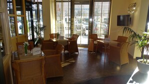 Un vestíbulo de hotel con mobiliario de mimbre, una recepción y grandes ventanales con persianas.