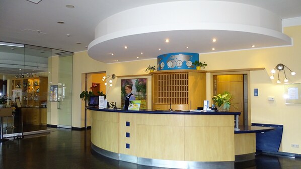 Zona de recepción de hotel con mostrador curvo, una mesa de recepción y un reloj de pared.