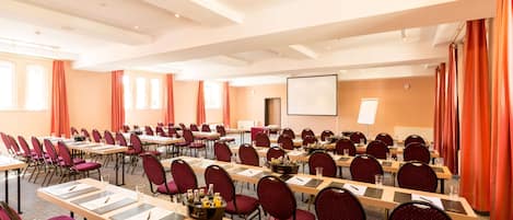 Un salón de conferencias con filas de mesas y sillas, una pantalla de proyector y una zona para presentaciones.