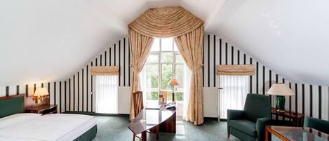 A hotel room with a large bed, a desk, two chairs, a window with curtains, and striped wallpaper.