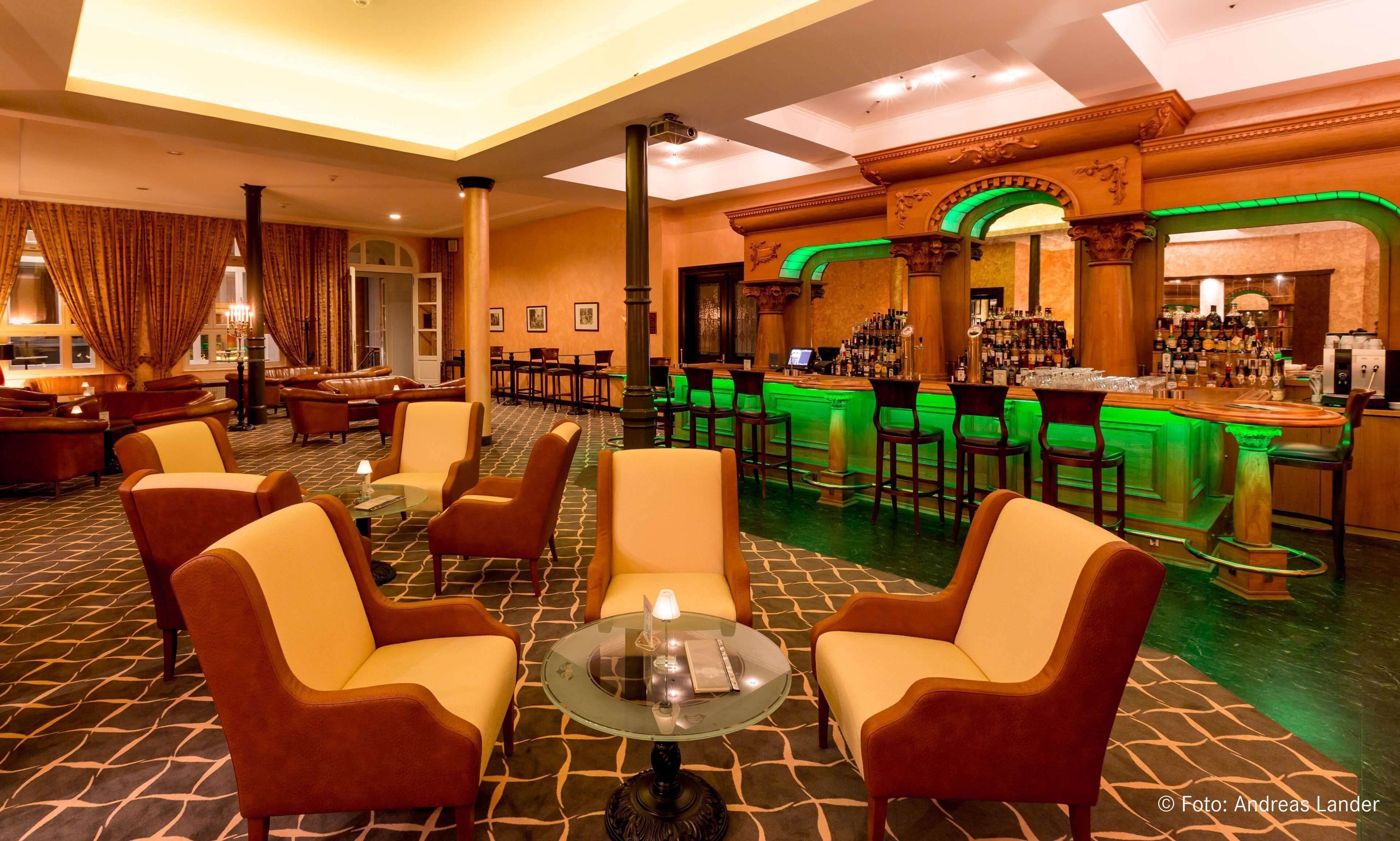 Un hall d’hôtel avec des canapés en cuir, un coin bar éclairé de lumière verte et un sol à motifs.