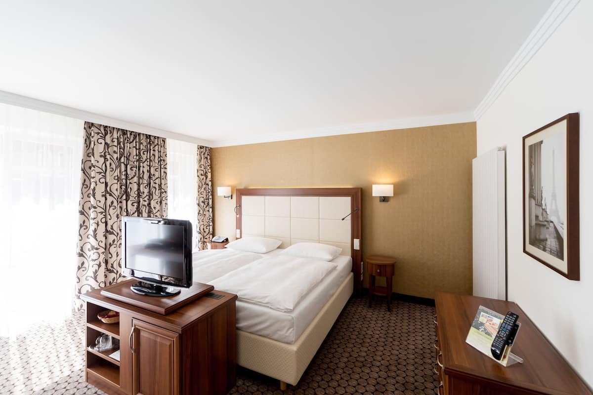 A hotel room with a bed, a TV on a wooden stand, a small table with a remote, and a framed picture on the wall.