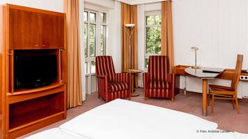 Habitación de hotel con cama, televisor sobre un soporte de madera, dos sillones a rayas, una mesita redonda pequeña, un escritorio con silla y ventanas grandes con cortinas.