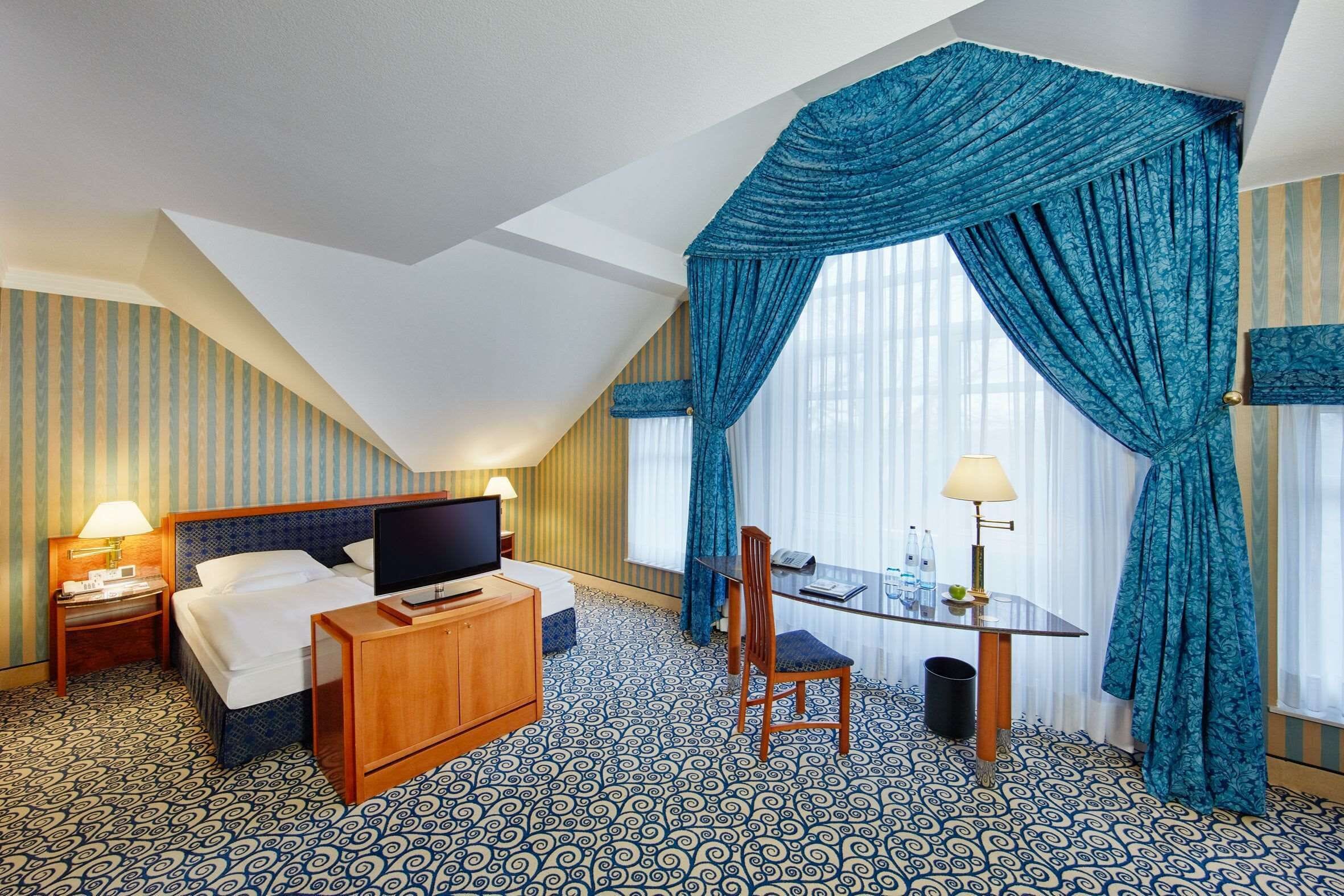 Habitación de hotel con cama, televisor, escritorio con silla y ventana grande con cortinas.