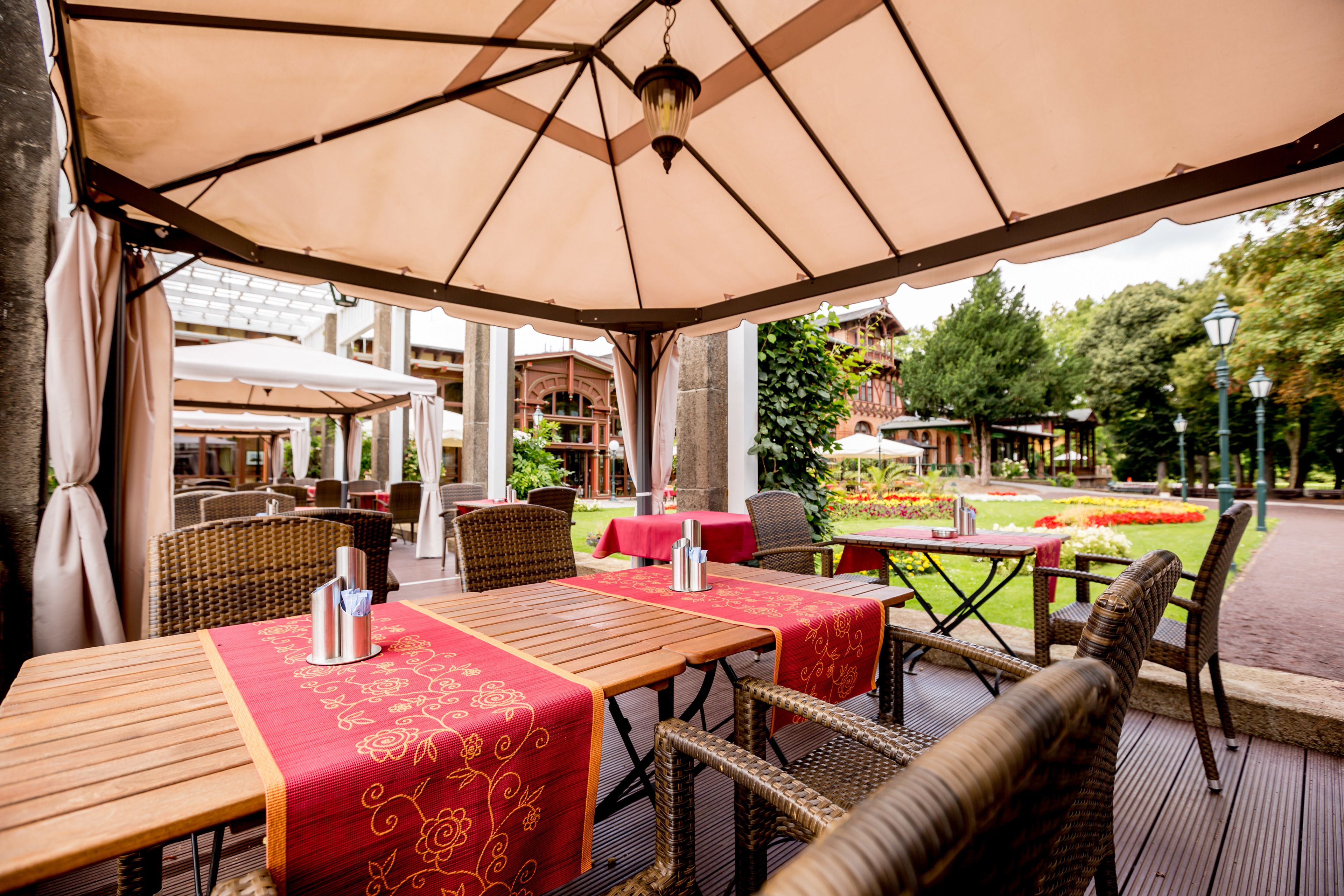 An outdoor dining area with tables covered by beige umbrellas, wicker chairs, and red tablecloths.