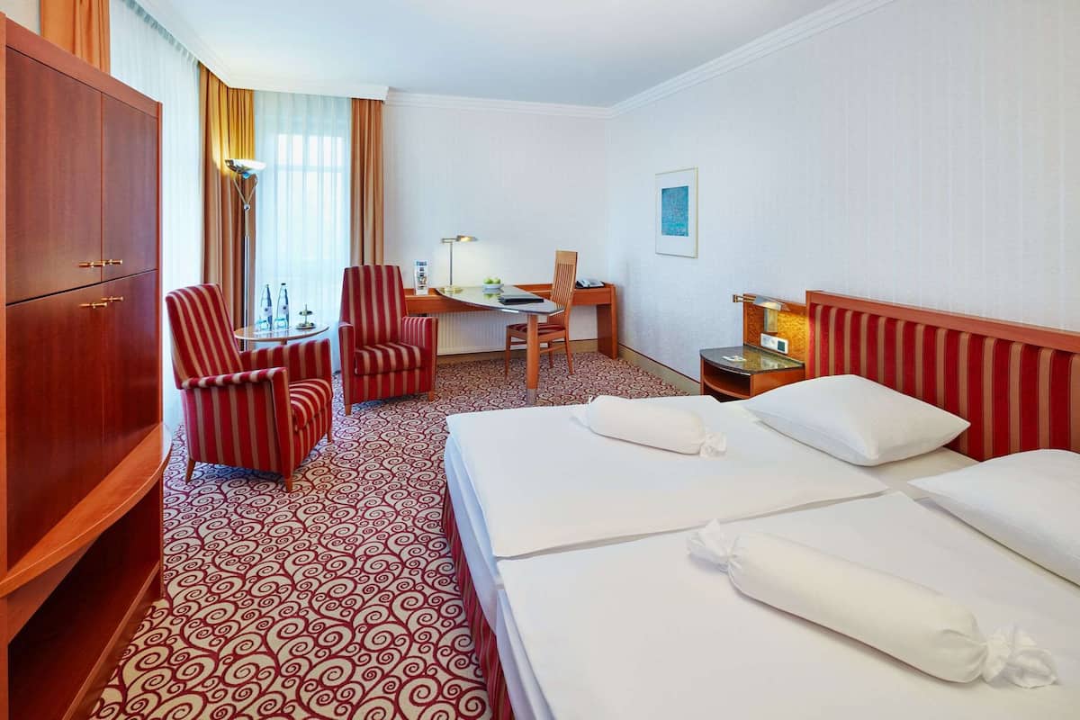 A hotel room with a large bed, a desk with a chair, a wardrobe, and a patterned carpet.