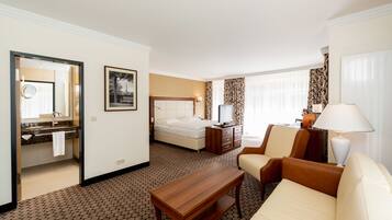 Habitación de hotel con cama, escritorio, sillón, sofá, televisión, lámpara, baño con espejo e imagen en la pared.