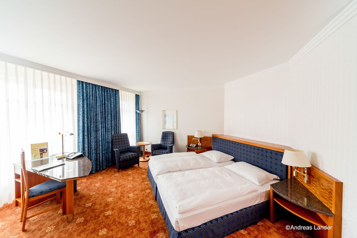 A hotel room with a large bed, a desk with a chair, a small table, and a lamp.