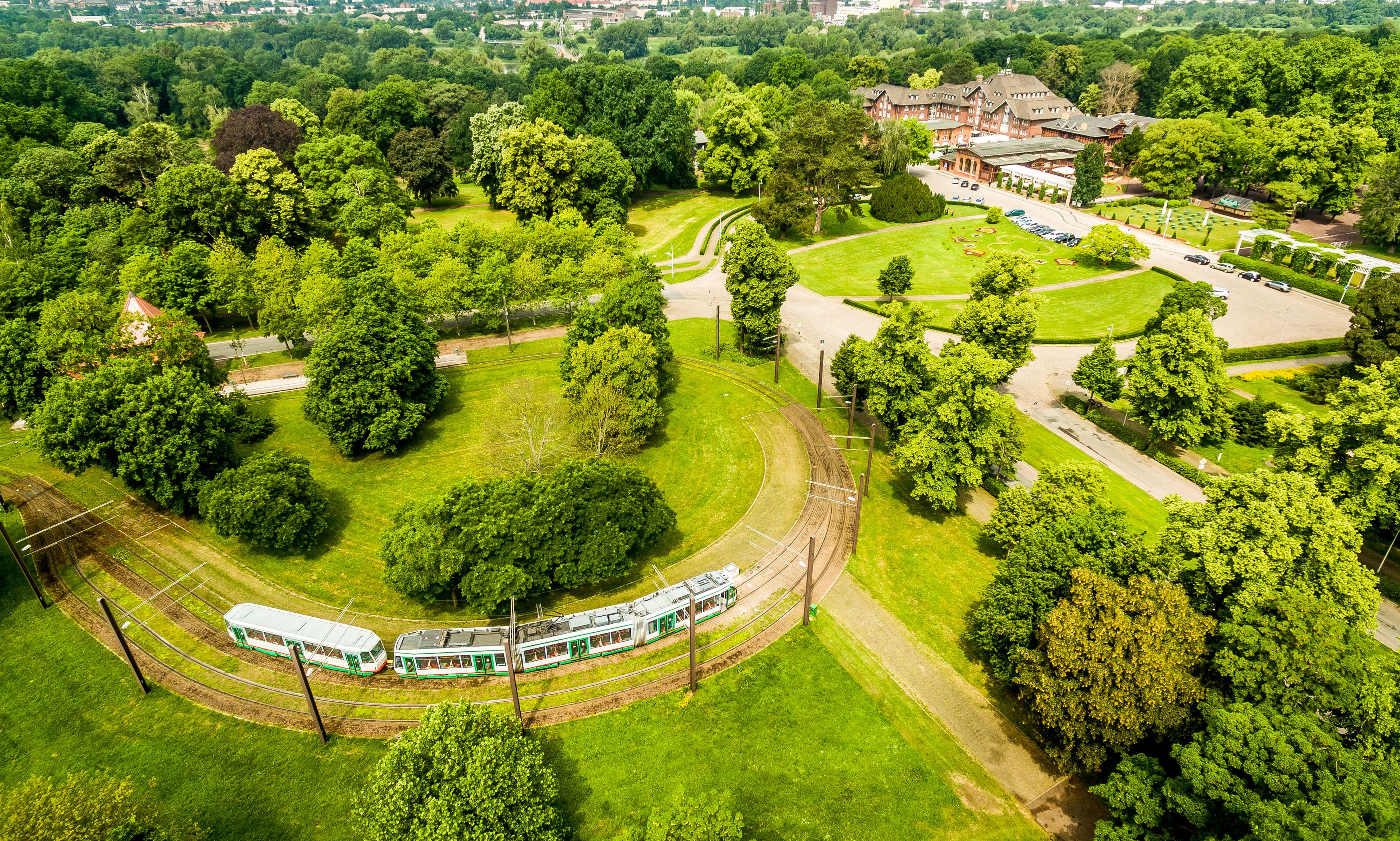 Vista aérea de un parque con un tranvía verde, árboles frondosos y un edificio con techo rojo.