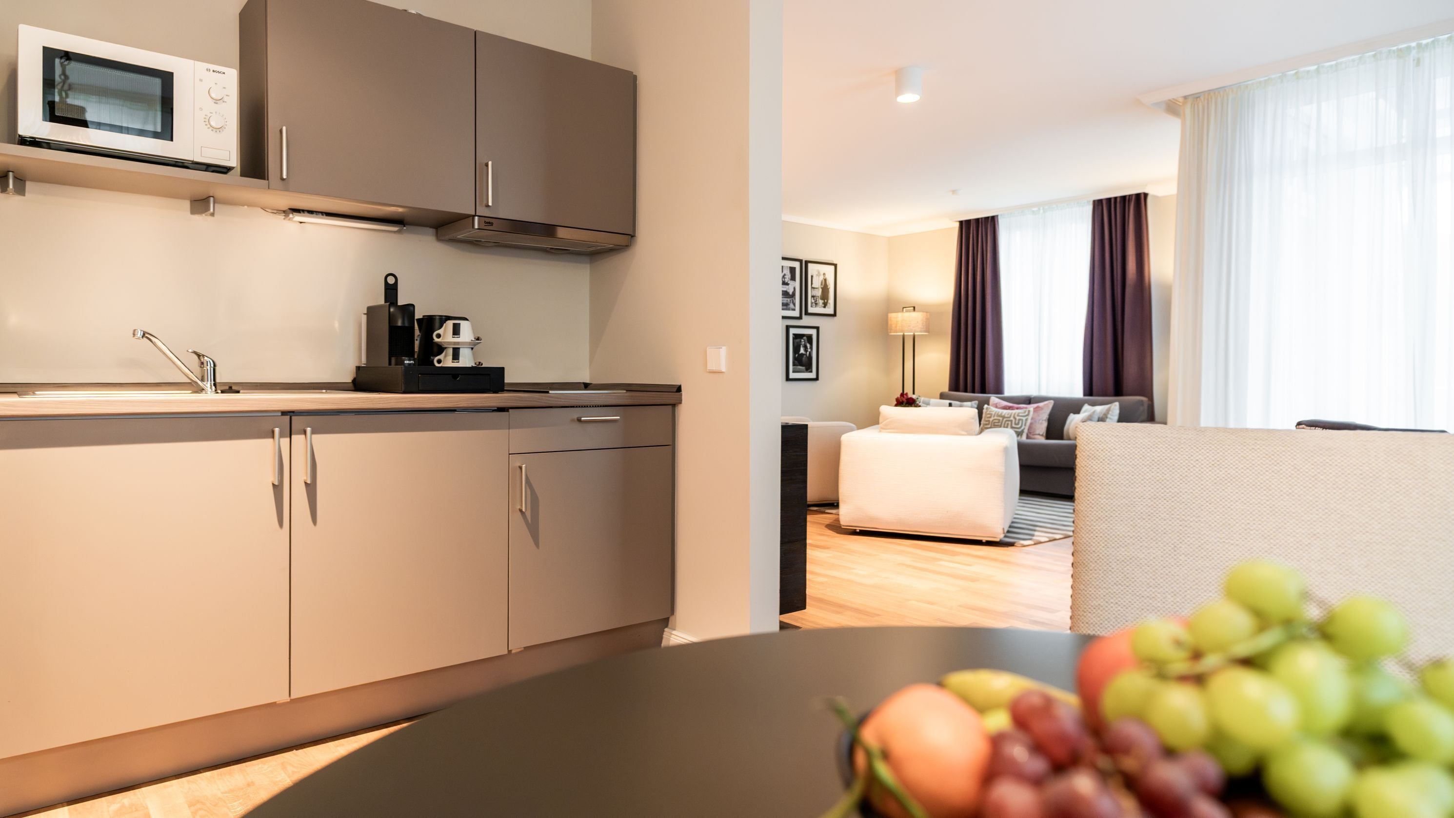 A modern kitchen with a microwave, coffee maker, and cabinets. There is a dining area with a table and a fruit bowl.