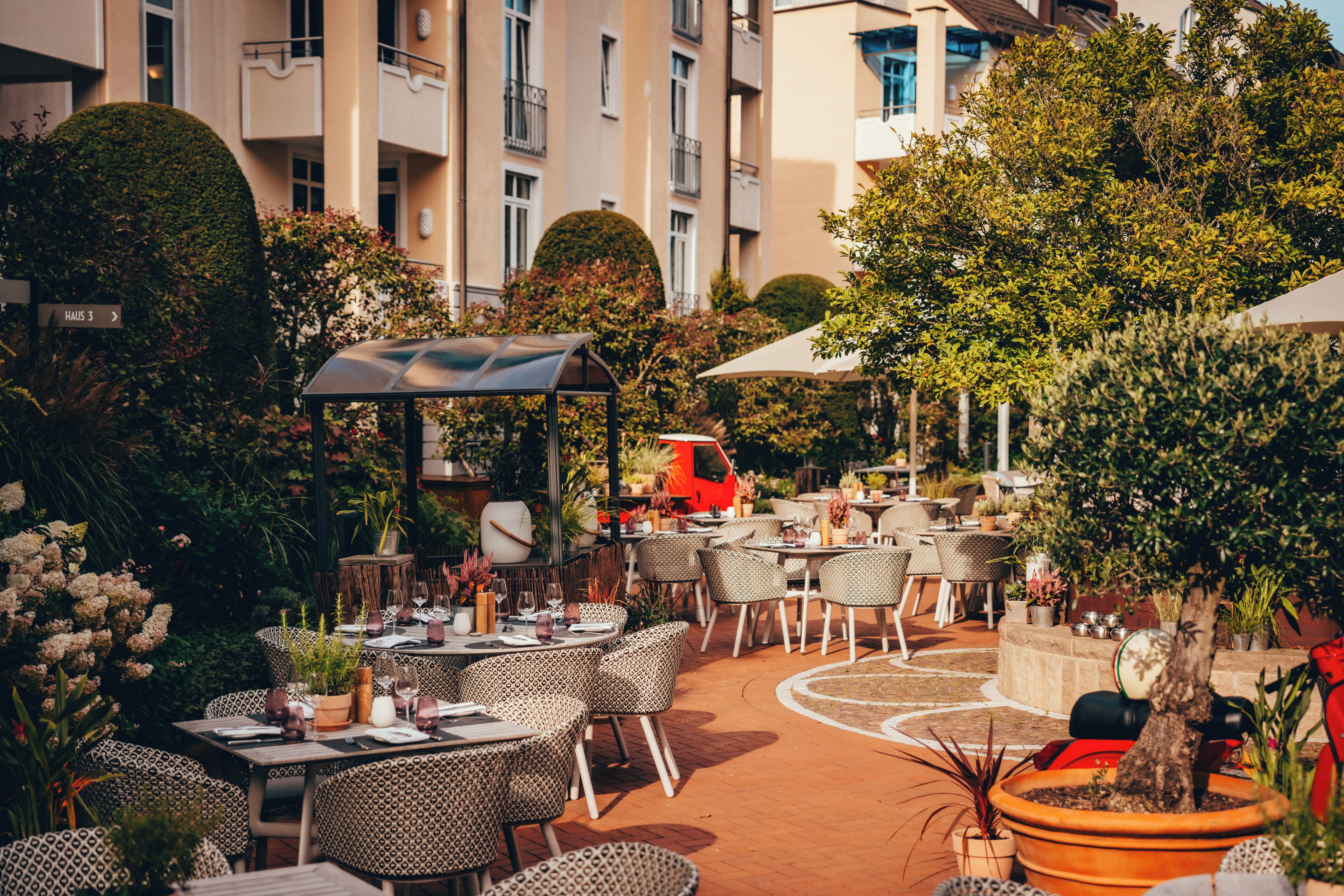 An outdoor dining area with tables and chairs, surrounded by greenery and a building in the background.