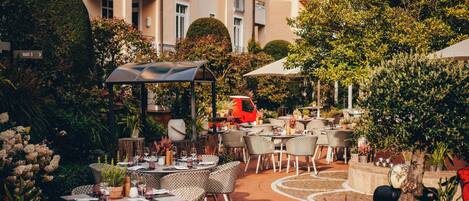 An outdoor dining area with tables and chairs, surrounded by greenery and a building in the background.