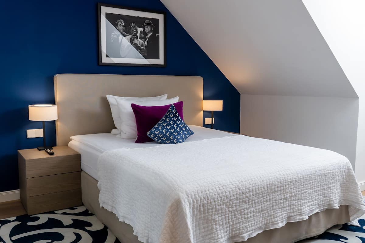 A neatly made bed with a white quilt, a purple pillow, and a blue patterned pillow. There are two bedside lamps on either side of the bed.