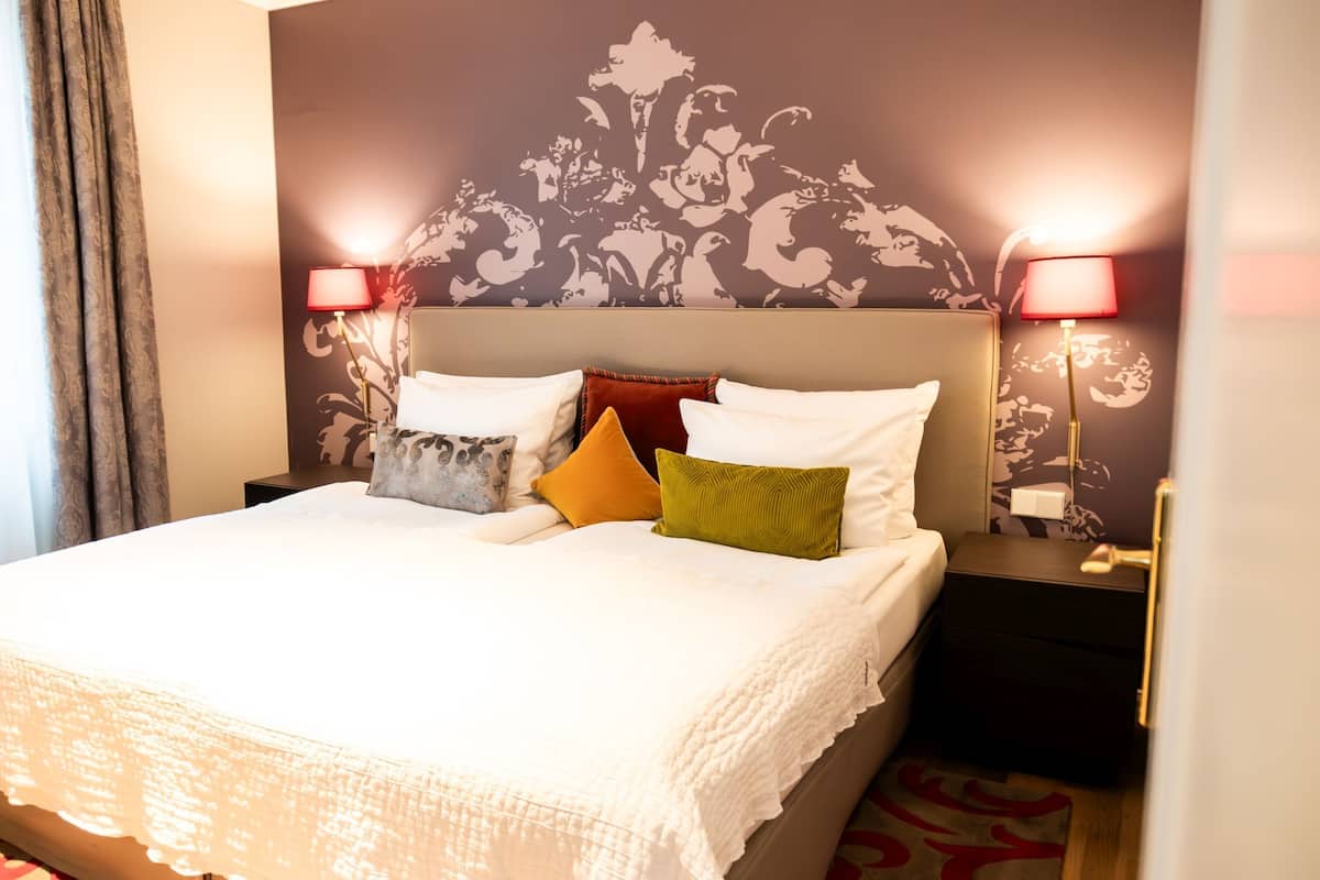 A hotel room with a large bed, two bedside tables, a headboard with a floral pattern, and two lamps on the nightstands.