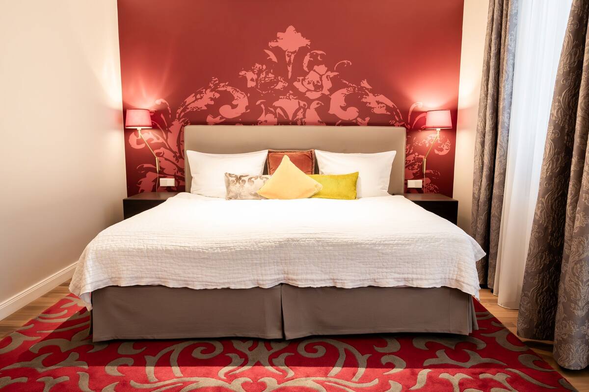 A neatly made bed with a white duvet, a brown headboard, and multiple pillows. There are two bedside lamps with red shades, a patterned red wall behind the bed, and a red and gold patterned carpet.