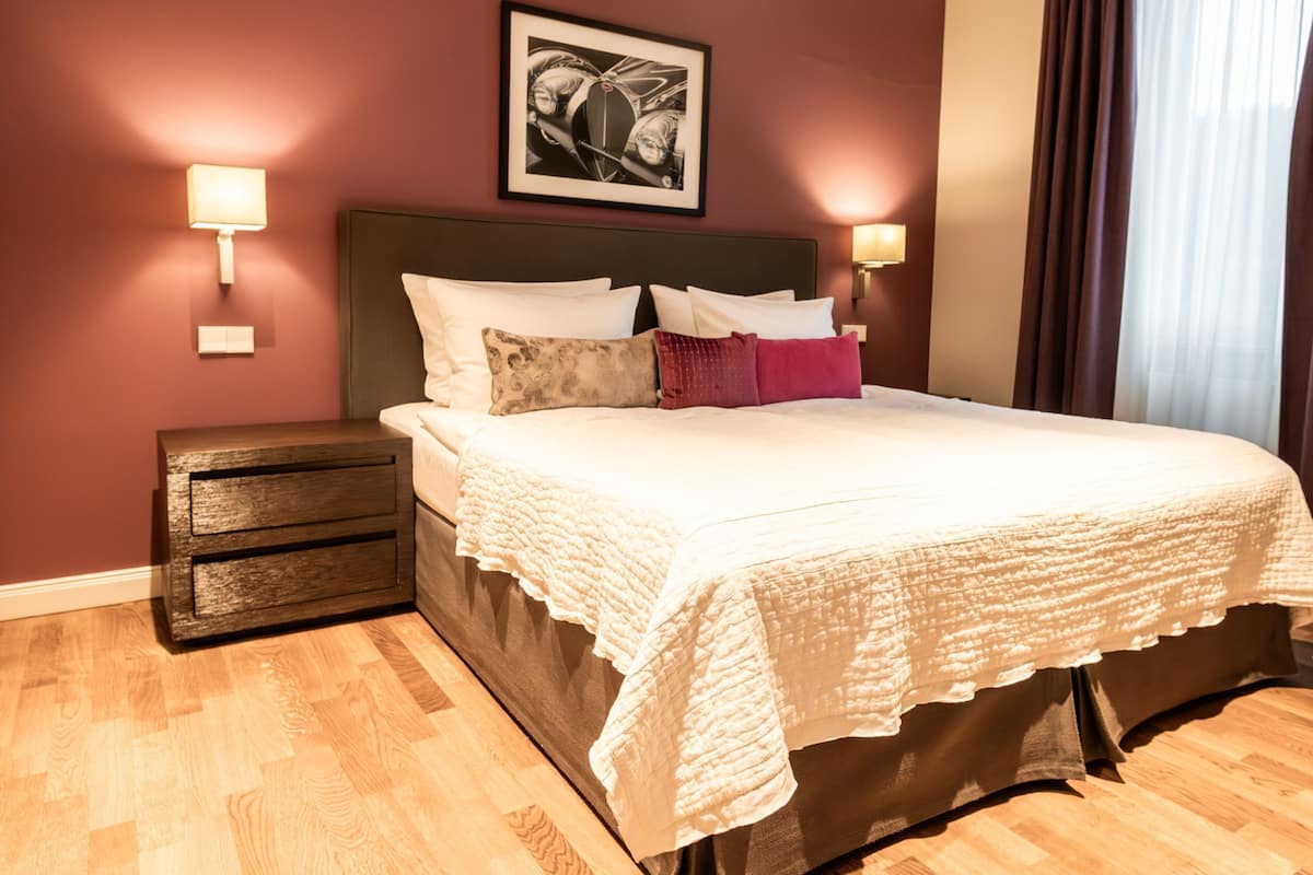 A bedroom with a bed, nightstands, a drawer unit, a framed picture on the wall, and a window with curtains.