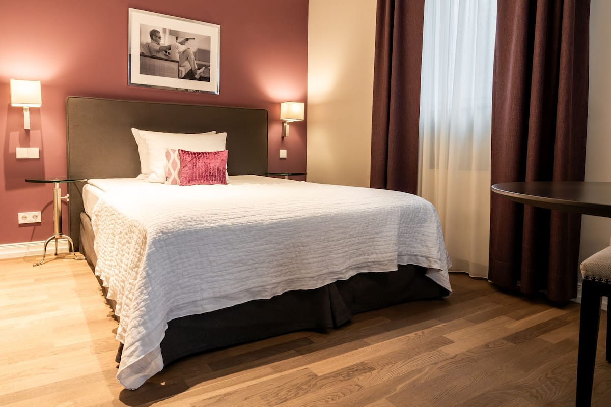 A neatly made bed with a white duvet and a pink accent pillow, a bedside table with a lamp, and a framed picture on the wall.
