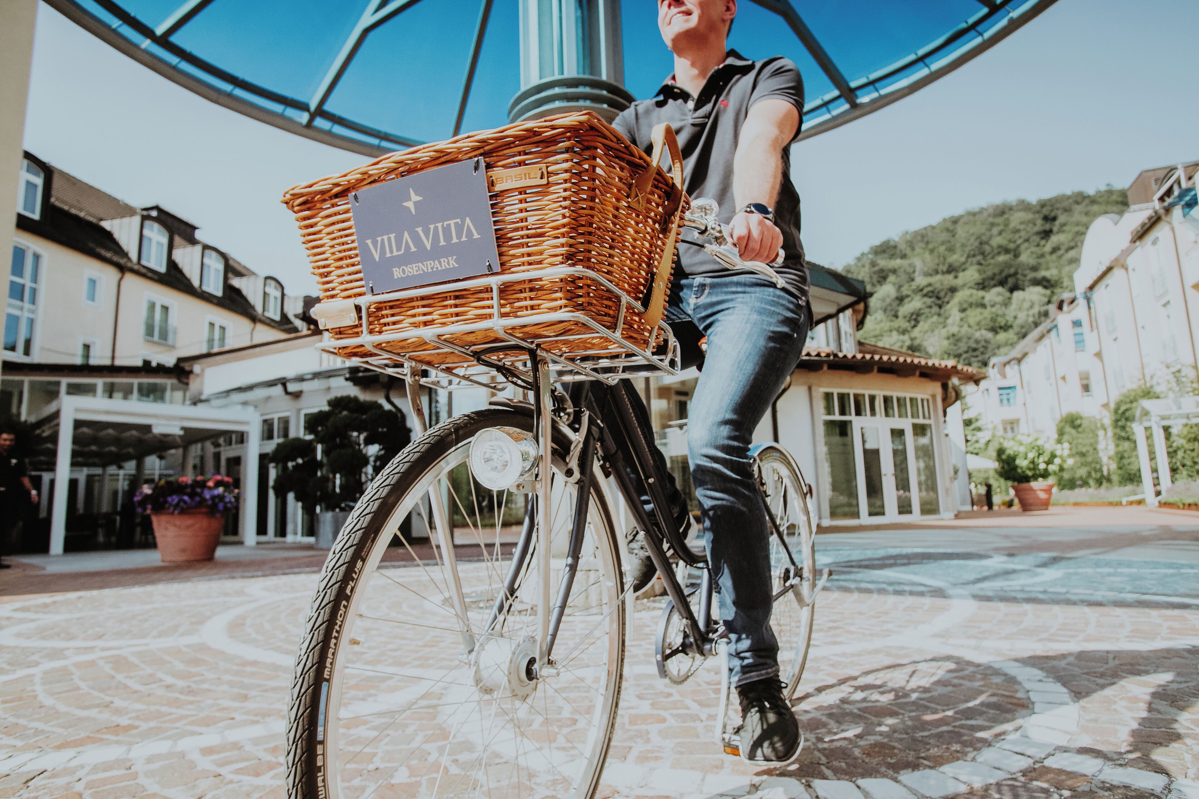 A person on a bicycle with a basket labeled 'VILLA ROSENMARK' in a courtyard with buildings and a hill in the background.