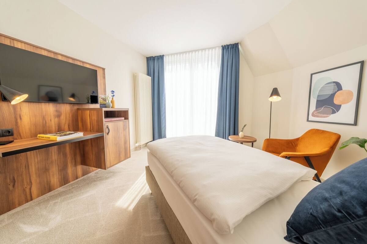 Ein Hotelzimmer mit einem Bett, einem Schreibtisch mit Lampe, einem Sessel, einem Heizkörper und einem Fenster mit Vorhängen.