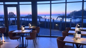 Restaurante com mesas e cadeiras de madeira, amplas janelas com vista para a orla.
