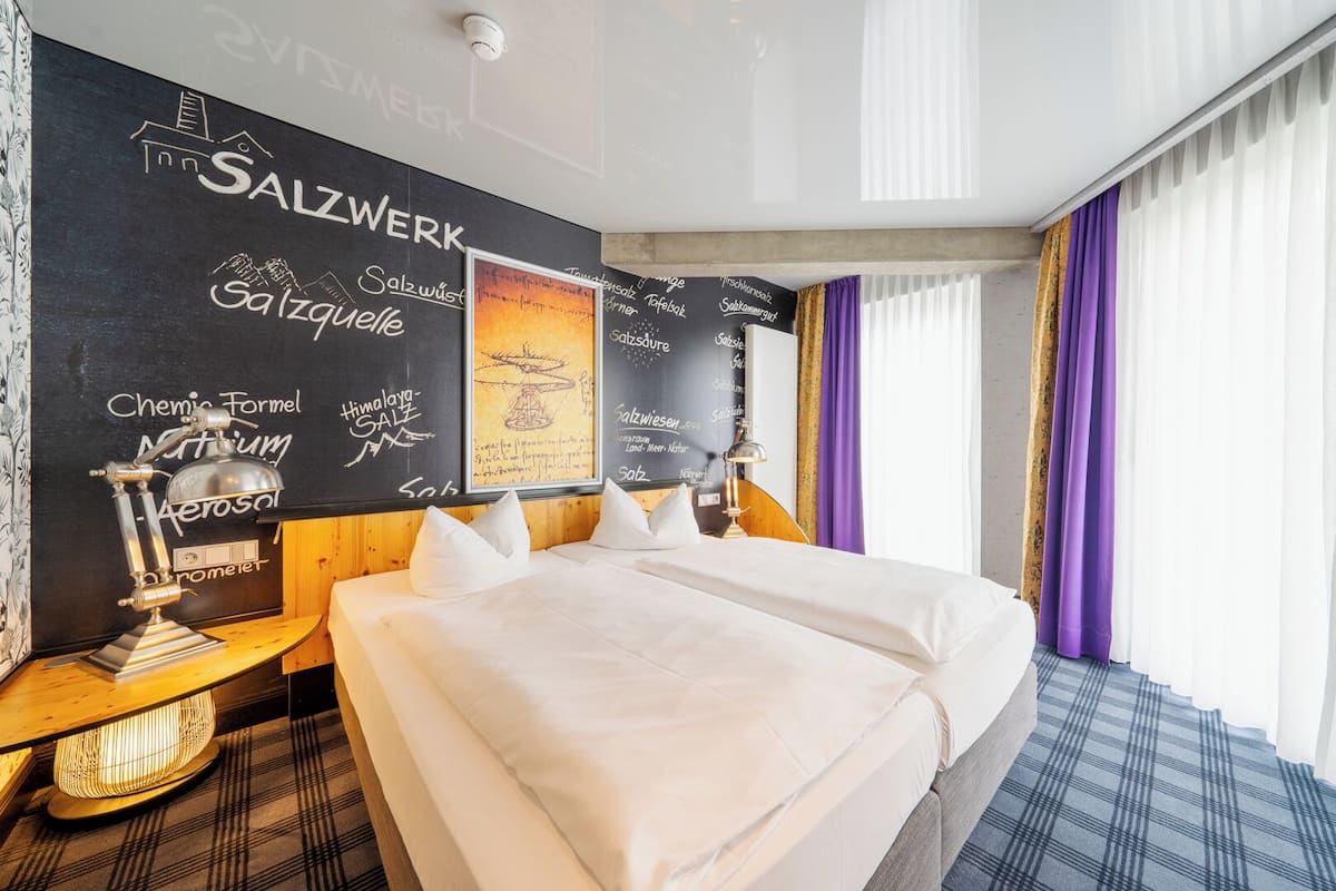 Ein Hotelzimmer mit einem großen Bett, einem Nachttisch, einer Wandleuchte und einem Fenster mit violetten Vorhängen.