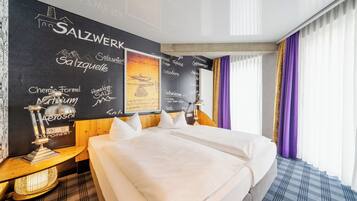Una habitación de hotel con una cama grande, una mesita de noche, una lámpara de pared y un ventanal con cortinas moradas.