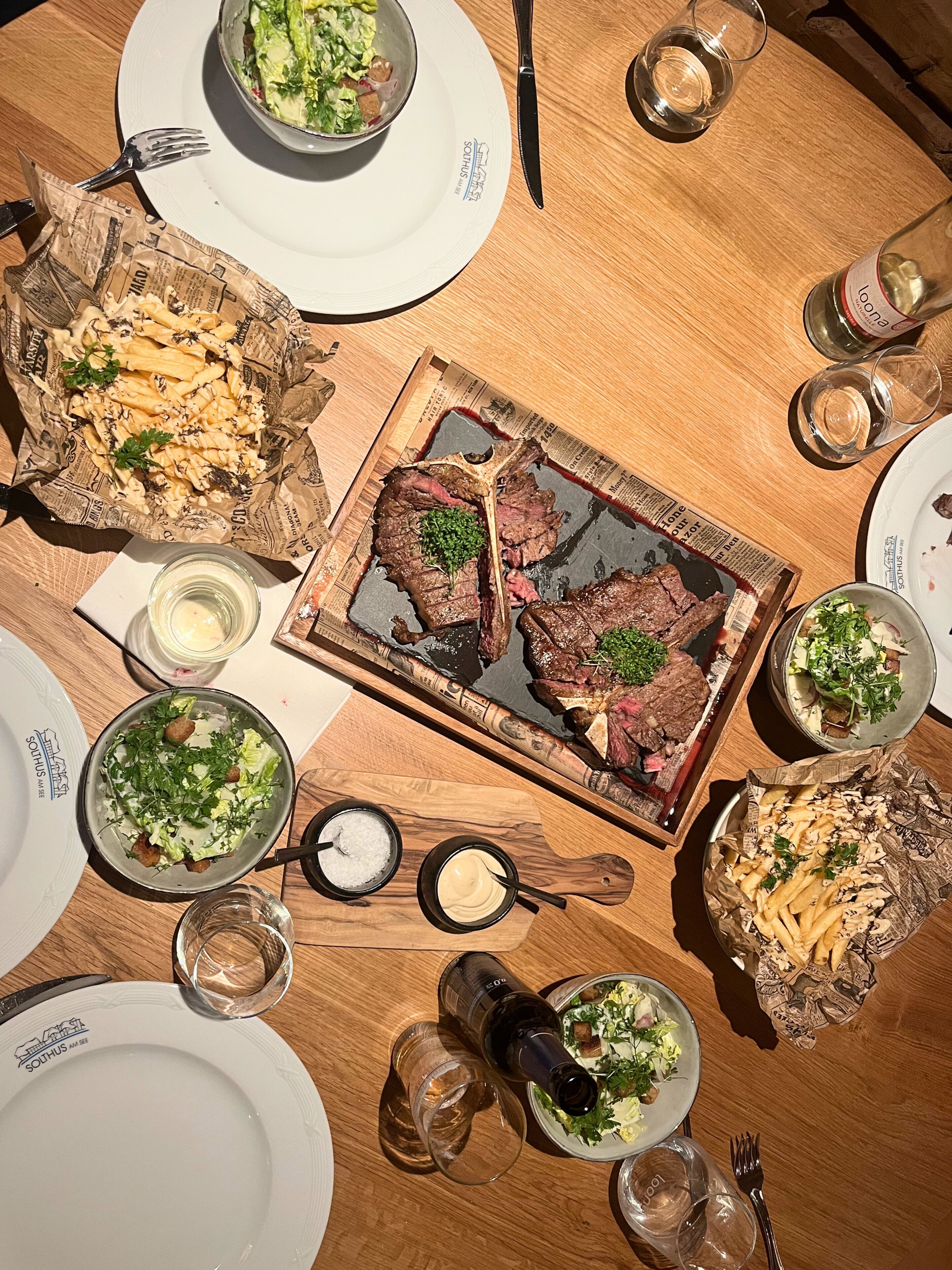 Una mesa redonda de madera con una variedad de platos, incluyendo carne, pasta y ensaladas, acompañada de copas de vino y cubiertos.