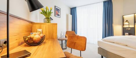 A hotel room with a wooden desk, a chair, a bed, a lamp, and a window with curtains.