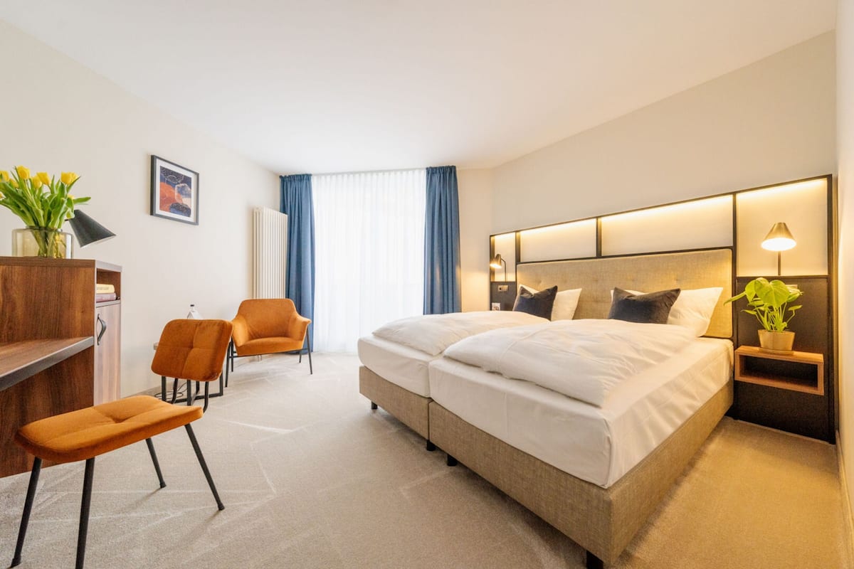 Ein Hotelzimmer mit einem großen Bett, einem Schreibtisch mit Stuhl, einem kleinen Tisch mit Lampe und einer Topfpflanze.