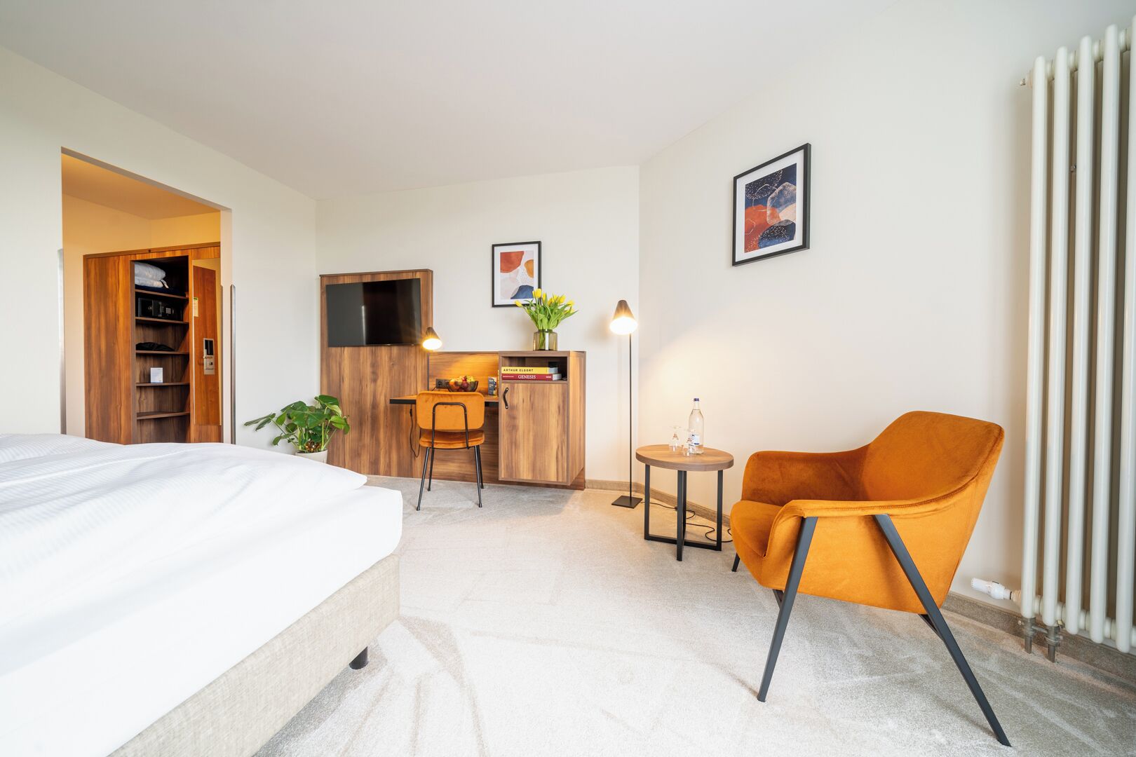 A hotel room with a bed, a desk with a chair, a small table with a lamp, and a radiator.
