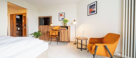 A hotel room with a bed, a desk with a chair, a small table with a lamp, and a radiator.